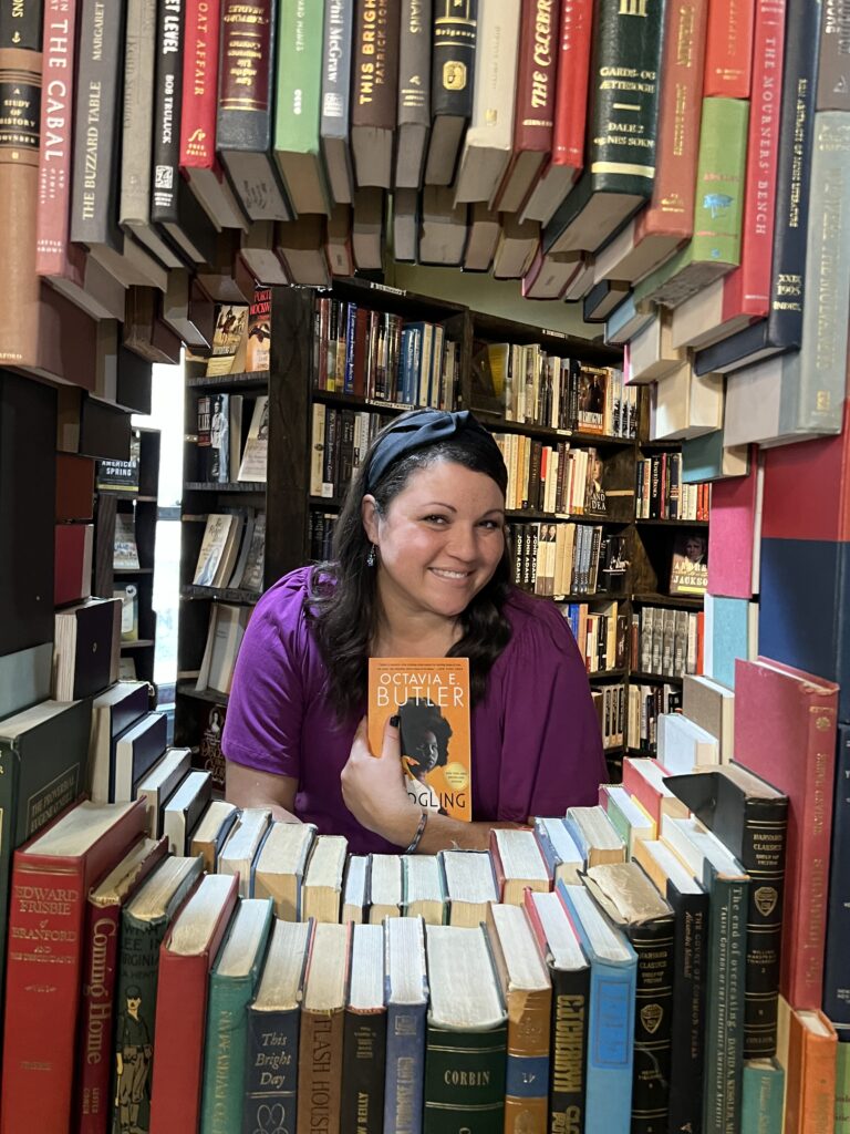 Renee is holding a book by Octavia Butler while peeking through a circle of books. Photo location: The Last Bookstore in Los Angeles, California, USA.